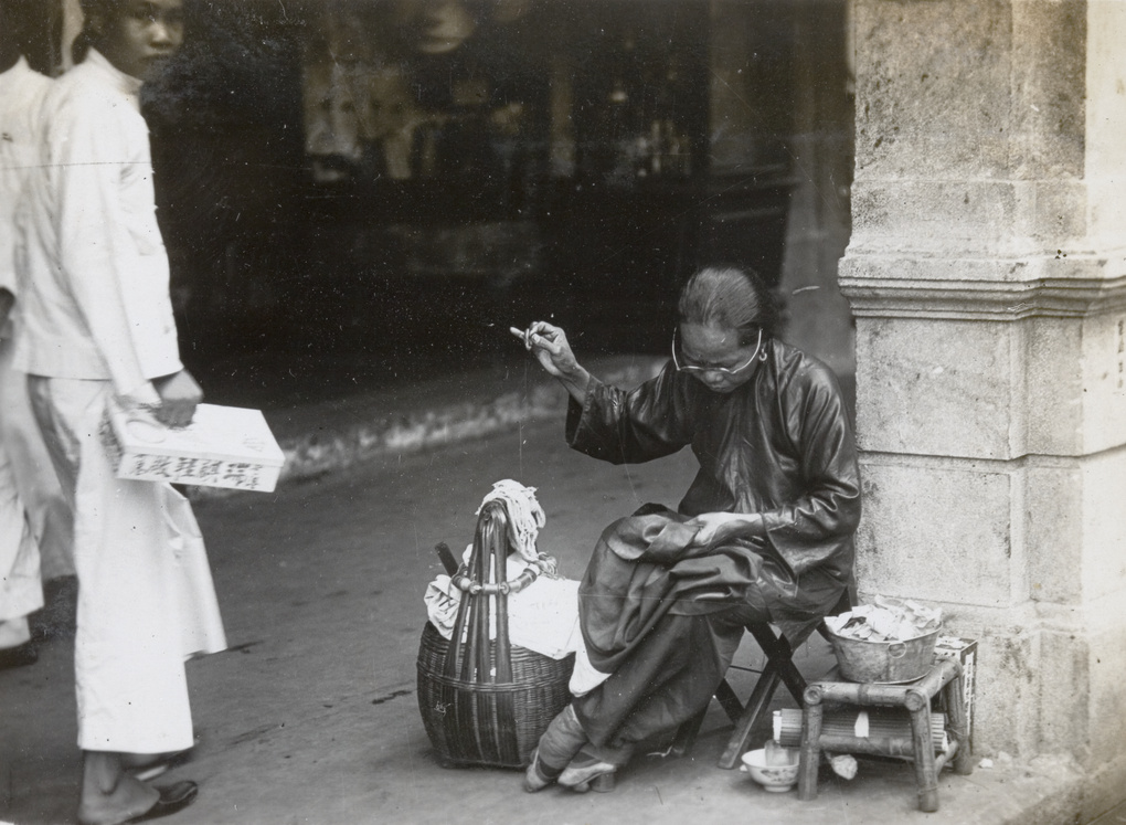 A seamstress with bound feet, repairing clothes by a shopping street, Hong Kong