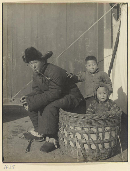 Man smoking a pipe, two children, and a basket near a vegetable market