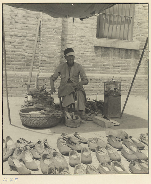 Shoe vendor seated with caged birds under a canopy next to a display of his wares