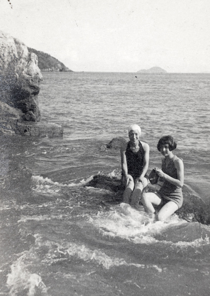 Maggie Henderson and Gladys Hutchinson, wearing bathing costumes and sitting on a rock in a bay, Hong Kong