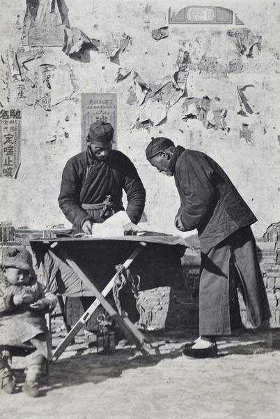 Child sitting beside the street stall of a letter writer with a customer