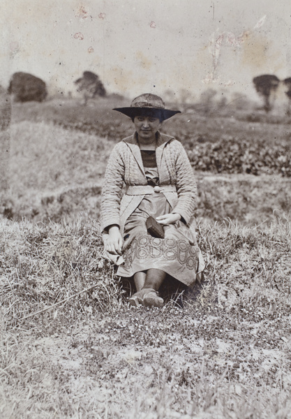 Sarah Hutchinson holding a camera and displaying the decorative needlework detail on her dress, sitting in a field near Tongshan Road, Hongkou, Shanghai