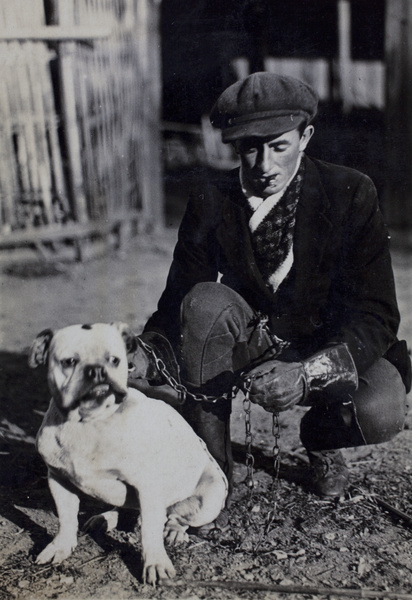 Unidentified man wearing a newsboy cap, protective gauntlets and smoking a cigar while posing with a bulldog on a chain leash, Kalgan Dairy Farm, Shanghai