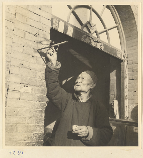 Man feeding a bird tethered to a perch beside a doorway with inscriptions in Baoding