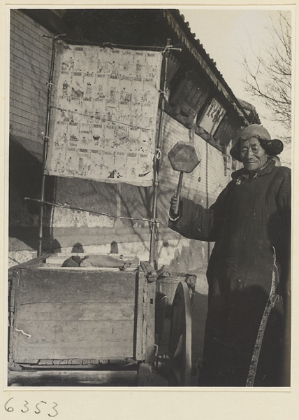 Tracery candy vendor holding a drum called a da tao and standing next to a cart with a sign showing candy designs