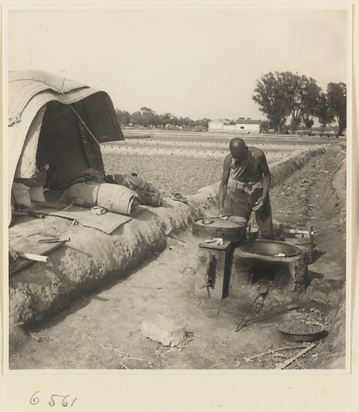 Man cooking at a stove in front of a mat shed next to fields