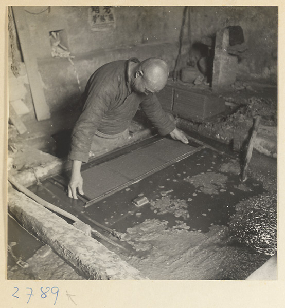 Man lifting a screen covered with paper pulp out of a vat in a paper-making shop