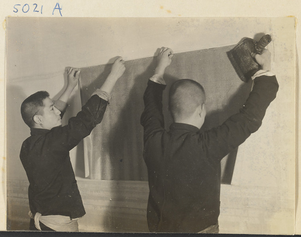 Interior of a scroll-mounting shop showing mounters attaching a backed scroll painting to a drying board called a zhuang ban