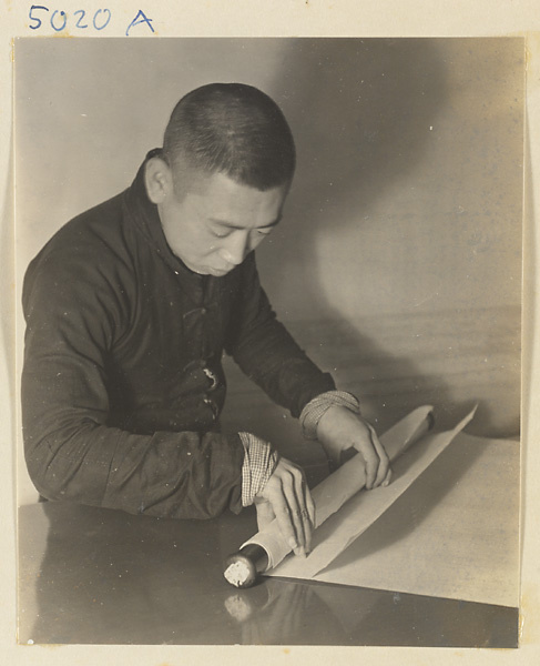 Interior of a scroll-mounting shop showing a man attaching the bottom roller bar called a zhou to a scroll painting