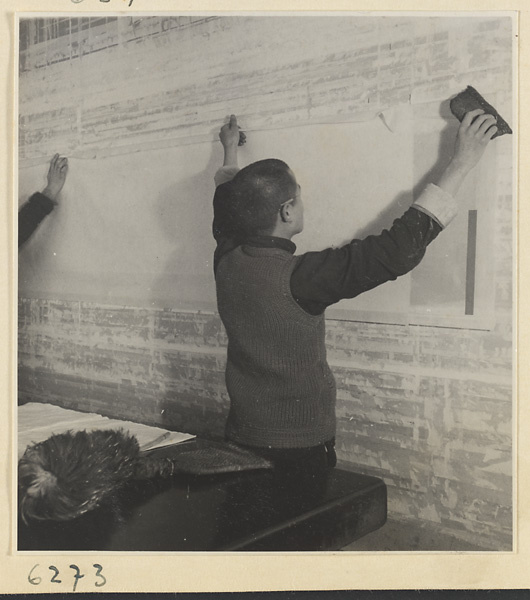Interior of a scroll-mounting shop showing two men hanging a scroll on a drying board called a zhuang ban