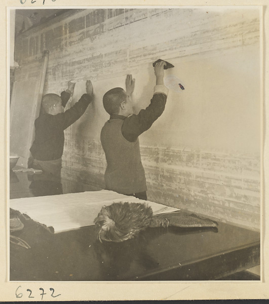 Interior of a scroll-mounting shop showing two men hanging a scroll on a drying board called a zhuang ban