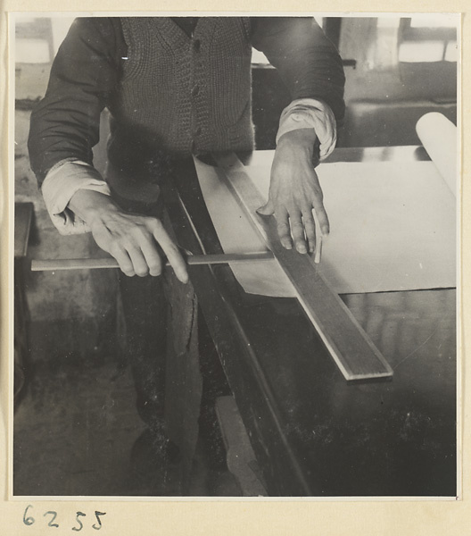 Interior of a scroll-mounting shop showing a man measuring the edge of a scroll painting