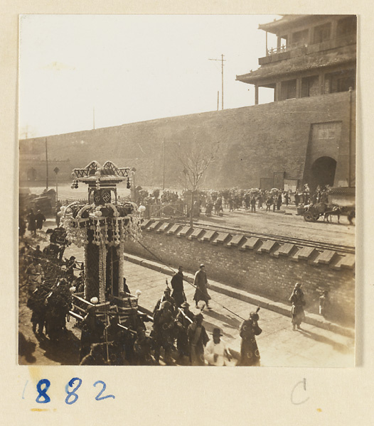 Funeral procession with a paper structure containing soul tablet and portrait of the deceased passing through a city gate