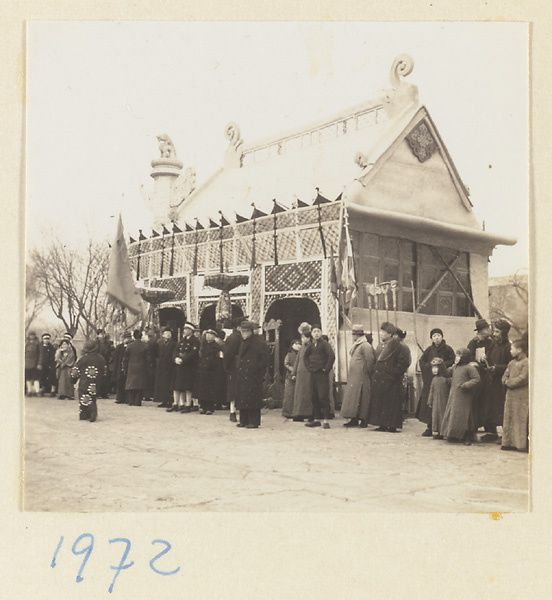 Members of a funeral procession holding flags, furled umbrellas, and staffs outside a paper pavilion erected for the funeral