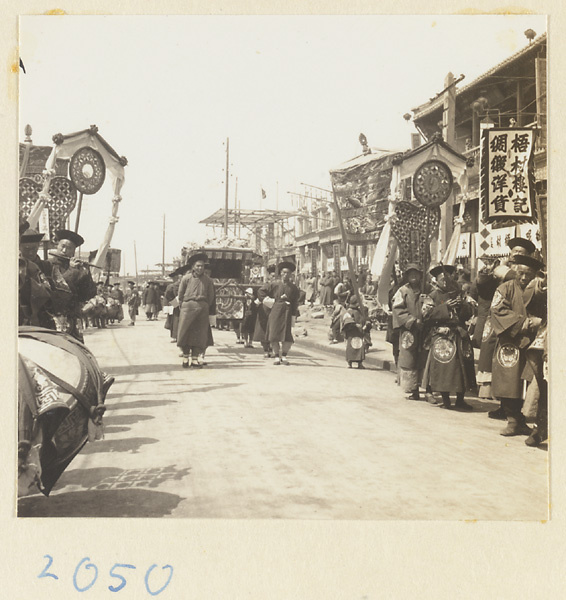 Members of a wedding procession playing musical instruments and carrying draped mirrors, fan-shaped screens, umbrellas, and sedan chair past shops