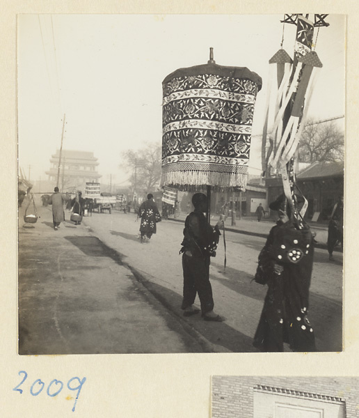 Members of a funeral procession carrying umbrellas and banner near city gate