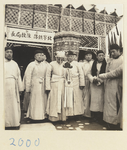 Principal mourner in white cap holding the soul banner and other mourners standing in front of a paper pavilion erected for the funeral