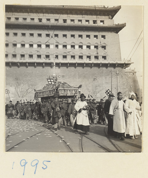 Principal mourner wearing white cap, man with drum keeping a beat for coffin bearers, and draped coffin being carried past Zhengyangmen (正阳门) / Qianmen (前门), Beijing