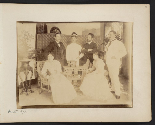 Group of four unidentified men standing around two young women sitting at small rattan table inside house in Canton