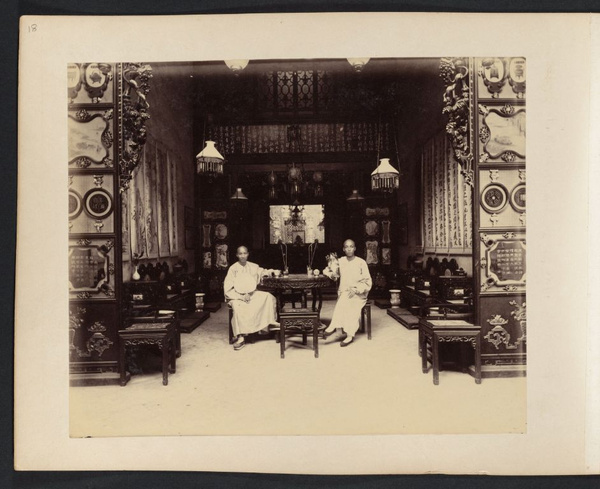 Two young Chinese men sitting at table in room with carved wood furniture and partitions, Canton