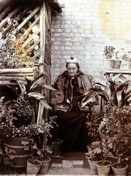 A woman wearing a fur-lined silk Chinese jacket, sitting among potted plants