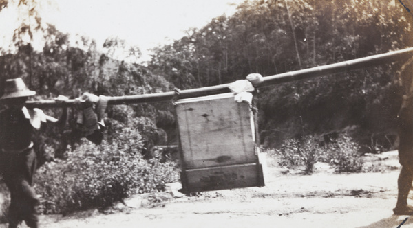 Two porters carrying a wooden crate on a long bamboo pole