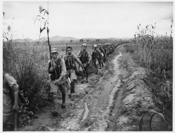 Soldiers (in vanguard) walking  along a track beside fields with crops, 1938