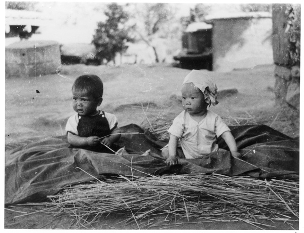 Yinhua, the baby son of Guo Qinglan and Dr Dwarkanath S. Kotnis, sitting on a sheet on top of some hay, with another baby, Jinchaji
