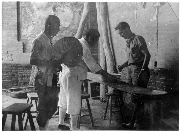 George E. Taylor with two Chinese men, one with a fan, next to a table, 1938