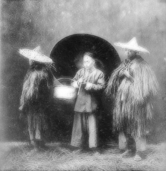 Three men posed in a photographer's studio – two wearing rice straw raincoats and bamboo hats