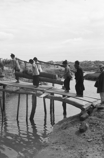 Four women carrying logs over a plank bridge