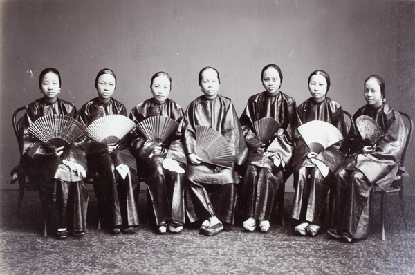 Seven women, with fans, sitting on bentwood chairs, in a photographer's studio