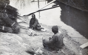 A woman splitting bamboo beside bundles of sticks on a river bank