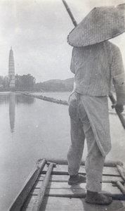 A boatman poling a sampan on a lake near a pagoda