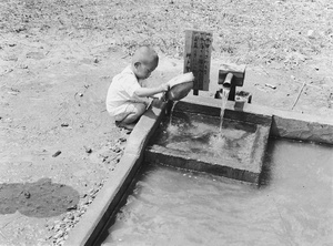 Boy using a straw hat to play with water flowing into a shallow concrete basin