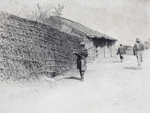 Man carrying a bundle of sticks passing a hedge on a country road