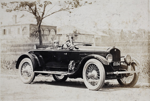 Baba Hansen posing behind the wheel of an automobile, with a uniformed driver in the front passenger seat, Dalney Road, Hongkou, Shanghai