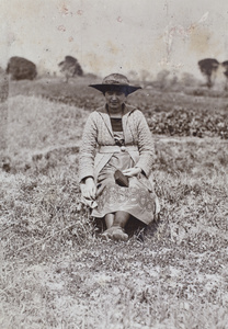 Sarah Hutchinson holding a camera and displaying the decorative needlework detail on her dress, sitting in a field near Tongshan Road, Hongkou, Shanghai