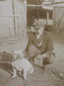 Unidentified man wearing a newsboy cap, protective gauntlets and smoking a cigar while posing with a bulldog on a chain leash, Kalgan Dairy Farm, Shanghai