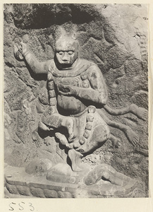 Animal-headed relief figure wearing necklace of skulls, carrying a body, and standing on a prone figure carved into the hillside at Yuquan Hill
