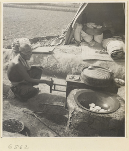 Man cooking at a stove in front of a mat shed next to fields