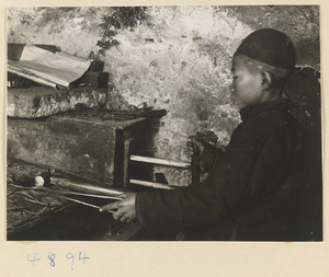 A boy operating a bellows for a forge in a workshop that makes iron pictures