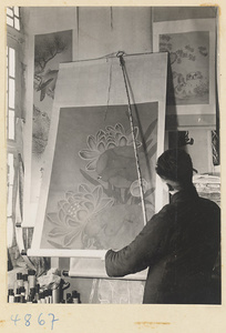 Man in a scroll-mounting shop using a scroll hanger called a hua cha to unroll a hanging scroll painting of flowers