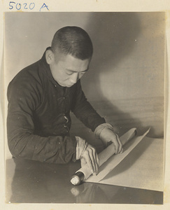 Interior of a scroll-mounting shop showing a man attaching the bottom roller bar called a zhou to a scroll painting