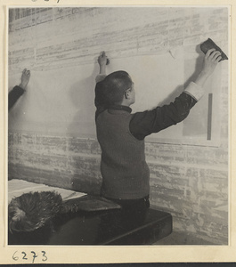 Interior of a scroll-mounting shop showing two men hanging a scroll on a drying board called a zhuang ban