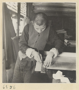 Interior of a scroll-mounting shop showing a man trimming the edge of a scroll painting with a steel paper knife