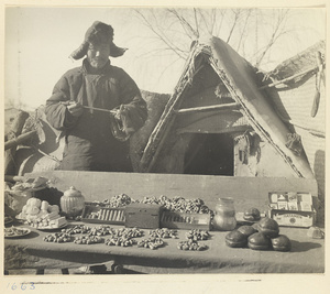 Street vendor with a pipe selling candy, peanuts, persimmons, and cigarettes in front of a mat shed