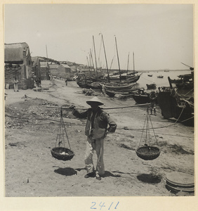 Man carrying baskets hung from shoulder pole on a beach in a fishing village on the Shandong coast