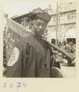Man with embroidered hat carrying gong and inscribed flag in wedding procession