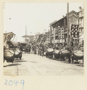 Members of a wedding procession playing drums and carrying draped mirrors, umbrella, and sedan chair past shops